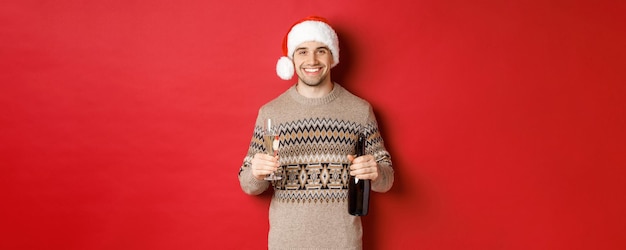 Concept of winter holidays, new year and celebration. Portrait of handsome man in santa hat and sweater, holding champagne and making toast on christmas party, standing over red background.