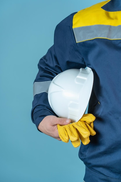 The concept of workplace safety. A male worker, an architect in work clothes holds a white protective helmet and gloves on a blue background in his hand.
