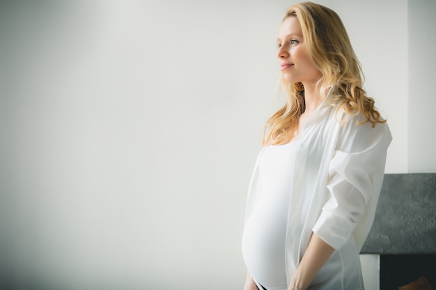 Concept - Young beautiful happy smiling pregnant caucasian blonde girl standing in white light living room