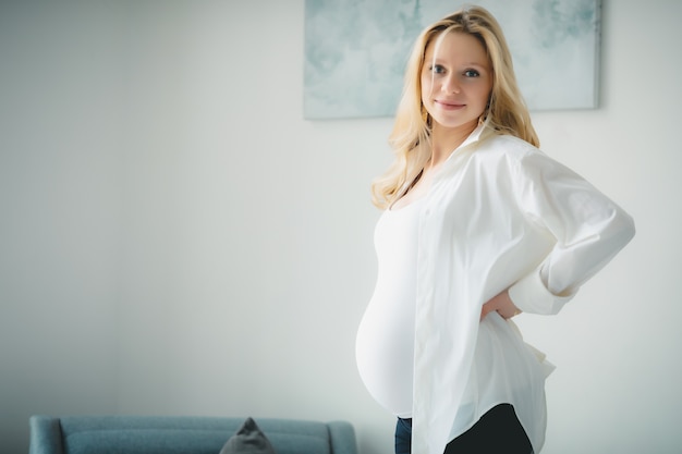 Concept - Young beautiful happy smiling pregnant caucasian blonde girl in white shirt standing in white light room