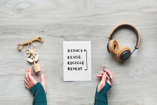 Concept zero waste conscious consumption flat lay above, Top view, hands holding trendy wooden flower, moleskin notebook with cover from craft cardboard, reading glasses and earphones on wooden table