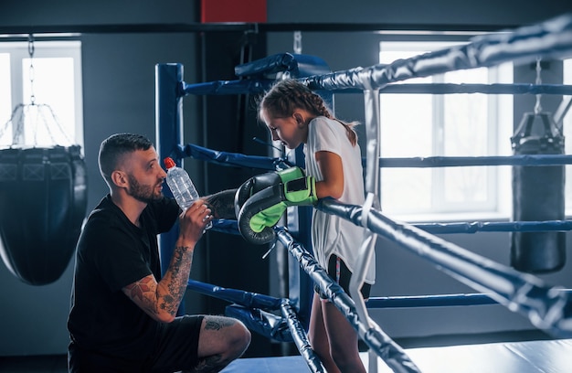 Conception of assistance. Young tattooed boxing coach teaches cute little girl in the gym.