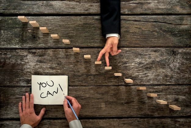 Conceptual image of personal growth and career development with businessman walking his fingers up wooden steps while his colleague or mentor encourages him with a You can message