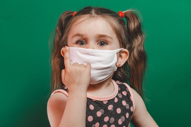 Conceptual photo of little girl in medical mask showing pointing finger on green background close up 2020