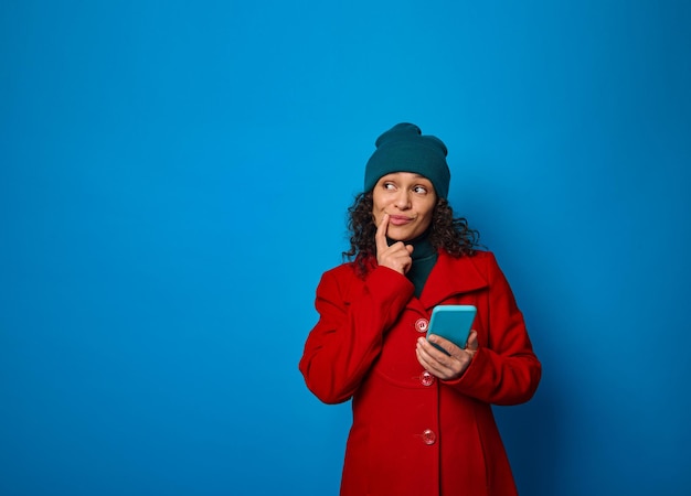 Conceptual portrait of a puzzled pensive pretty woman wearing bright red coat and warm green woolen hat holding smartphone and looking thoughtfully on a copy ad space , isolated on blue background