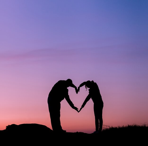 Conceptuele hartvorm, symbool van de mens. Vrouw en man hand silhouet over hemel bij zonsondergang, metafoor om lief te hebben.