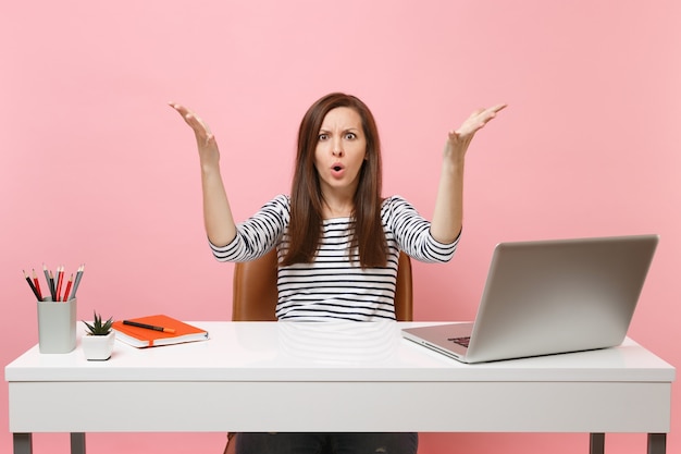 화가 난 여성은 파스텔 핑크색 배경에 격리된 현대적인 PC 노트북이 있는 흰색 책상에 앉아서 일을 하고 있습니다. 성취 비즈니스 경력 개념입니다. 공간을 복사합니다.