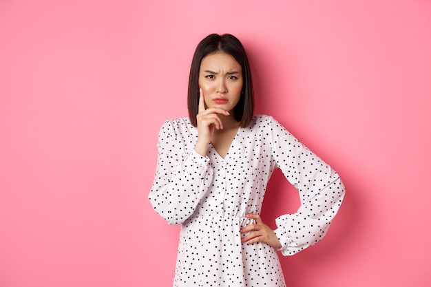 Concerned asian woman staring at camera suspicious, having doubts, thinking about something with serious face, standing against pink background