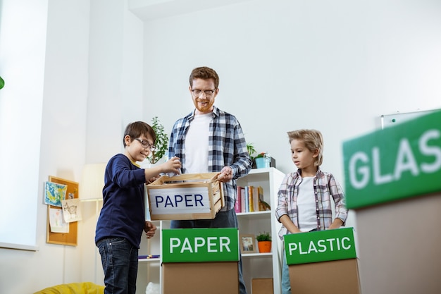 Concerned blonde boy. Ginger tall tutor working on sorting out different garbage while spending lessons with kids