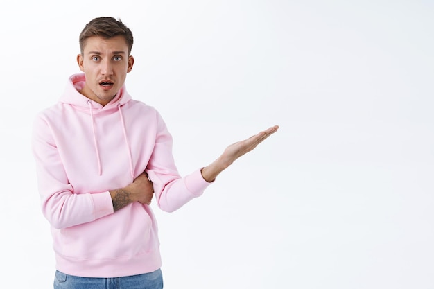 Concerned, frustrated blond young man in pink hoodie, pointing finger right at blank white space, staring camera with questioned complicated look