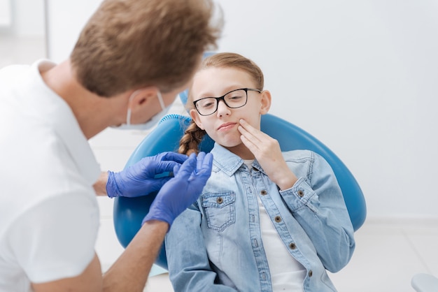 Concerned ginger teenage lady showing where the pain concentrating while paying a visit to her doctor and asking him for help