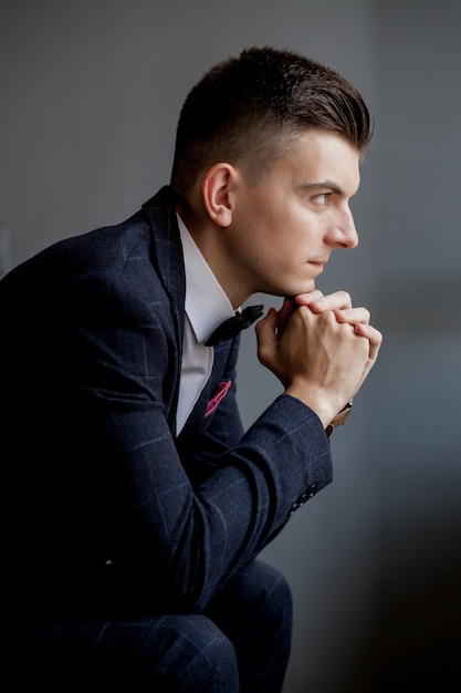 Concerned groom playing with his hands and looking to the side while wearing tuxedo, sitting on black studio background