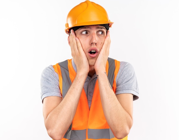 Concerned looking at side young builder man in uniform putting hands on cheeks