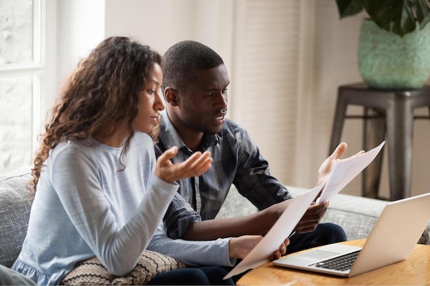 Concerned mixed race husband and wife take care of domestic bills reading papers at home worried black couple consider bank documents think of mortgage or loan check paperwork together