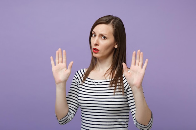 Concerned nervous young brunette woman in casual striped clothes posing isolated on violet purple background studio portrait. People lifestyle concept. Mock up copy space. Rising hands, showing palms.