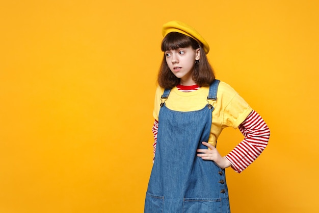 Concerned puzzled girl teenager in french beret, denim sundress looking aside, standing with arms akimbo isolated on yellow background. People sincere emotions, lifestyle concept. Mock up copy space.