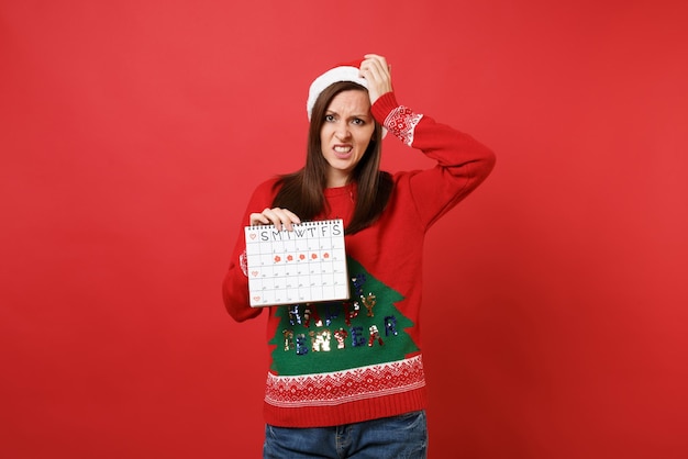 Concerned Santa girl holding female periods calendar for checking menstruation days isolated on red background. Medical healthcare gynecological. Happy New Year 2019 celebration holiday party concept.