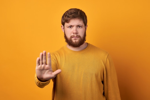 Concerned serious bearded man in round eyewear pulls palm towards camera