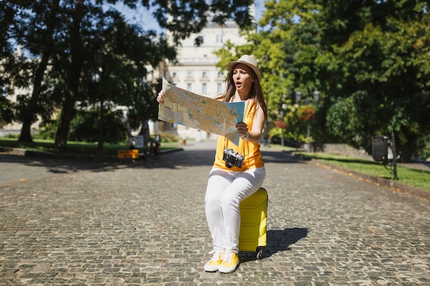 黄色い服の帽子をかぶった心配している旅行者の観光客の女性は、屋外の都市の都市地図検索ルートを探してスーツケースに座っています。週末の休暇で旅行するために海外旅行する女の子。観光の旅のライフスタイル。