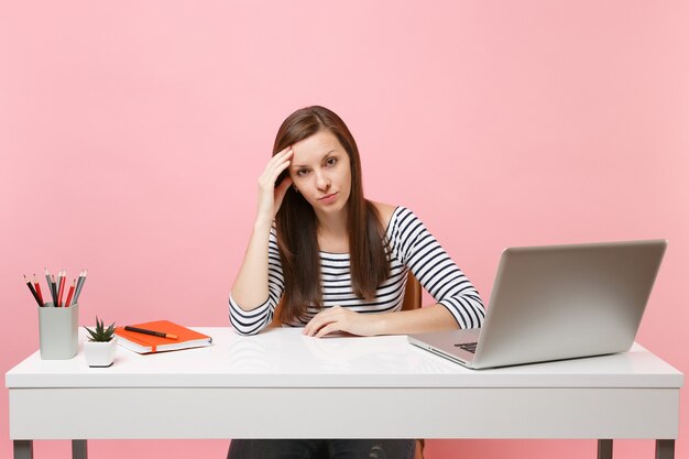 Concerned upset tired woman leaning on hand sit, work at white desk with contemporary pc laptop isolated on pastel pink background. Achievement business career concept. Copy space for advertisement.