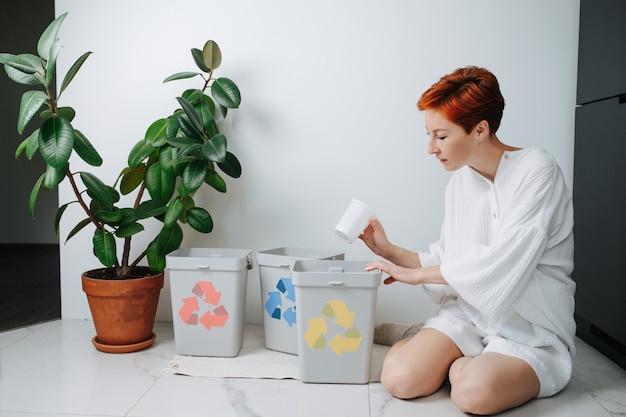 Concerned woman sitting on her legs, sorting garbage beween\
small recycle bins at home. they have differently colored arrows on\
them. holding plastic cup.