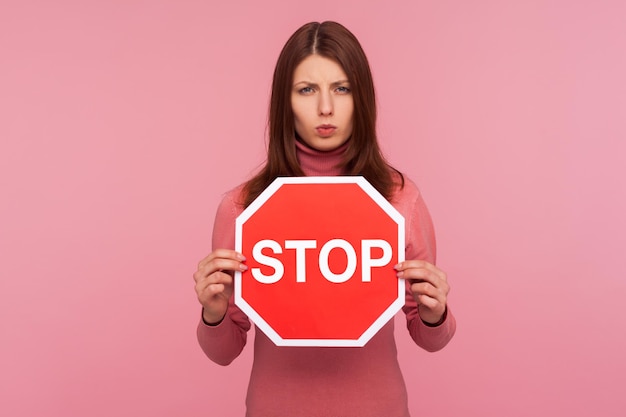 Concerned worried woman holding stop sign in hands warning\
about compliance with rules traffic laws indoor studio shot\
isolated on pink background