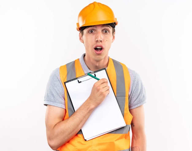 Concerned young builder man in uniform holding clipboard with pencil 