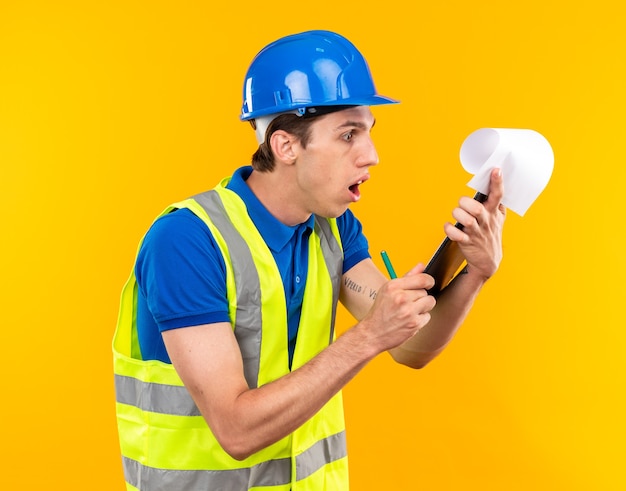 Concerned young builder man in uniform holding and looking at clipboard isolated on yellow wall