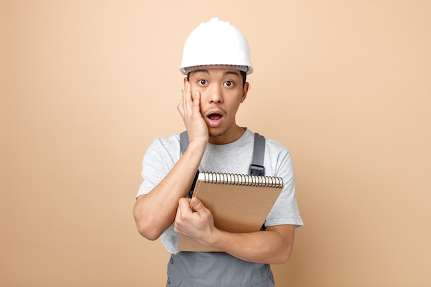 Concerned young construction worker wearing safety helmet and uniform holding notepad and pencil keeping hand on face 