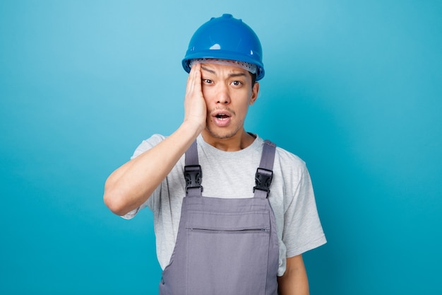 Concerned young construction worker wearing safety helmet and uniform keeping hand on head 