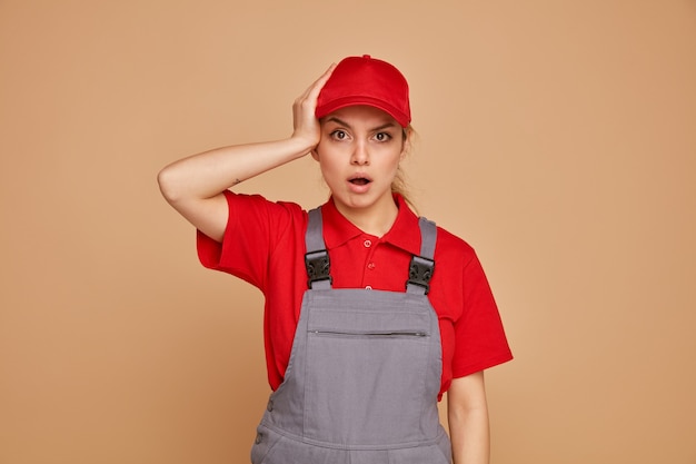 Concerned young female construction worker wearing uniform and cap keeping hand on head 