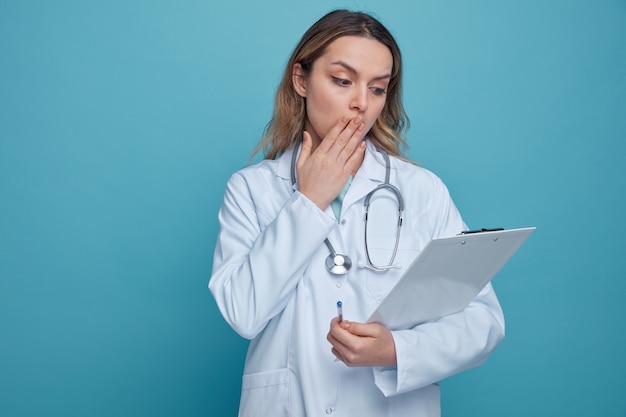 Preoccupato di giovani donne medico che indossa abito medico e stetoscopio intorno al collo tenendo la penna e appunti guardando negli appunti facendo oops gesto