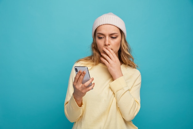 Concerned young girl wearing winter hat keeping hand on mouth holding and looking at mobile phone 