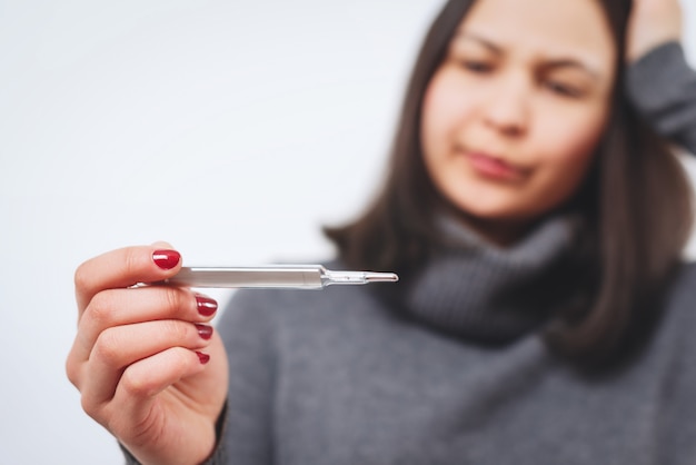 A concerned young woman in a gray sweater, holding a thermometer. Healthy lifestyle, treatment of patients with diseases, the concept of the cold season