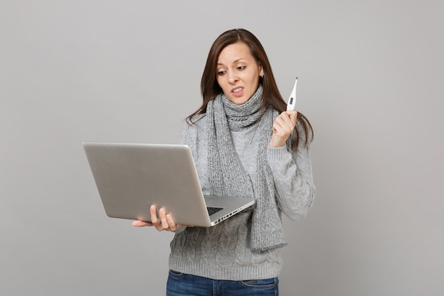 Giovane donna interessata in maglione grigio, sciarpa che lavora al computer pc portatile, tenendo il termometro isolato su sfondo grigio muro. stile di vita sano, consulenza sul trattamento online, concetto di stagione fredda.