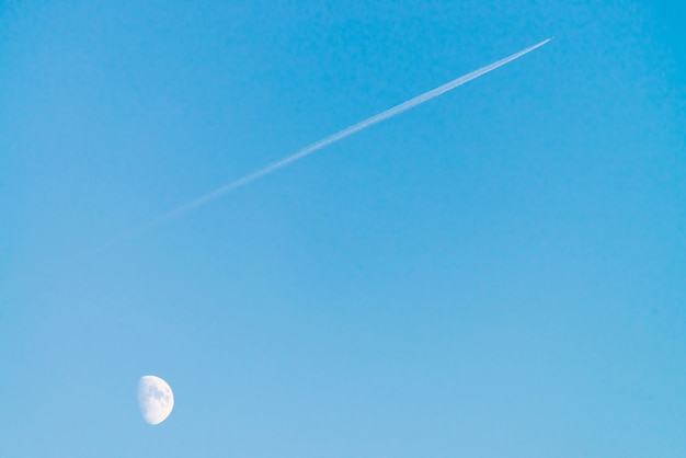 Condensation track of jet above moon in clear blue day sky. Minimalist blue background. Plane flies up diagonally.