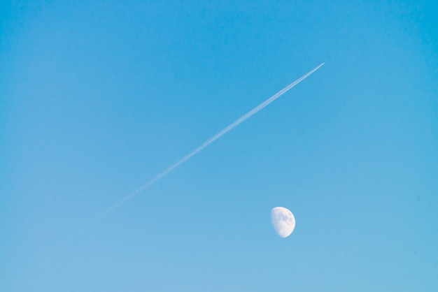 Condensation track of jet above moon in clear blue day sky.
