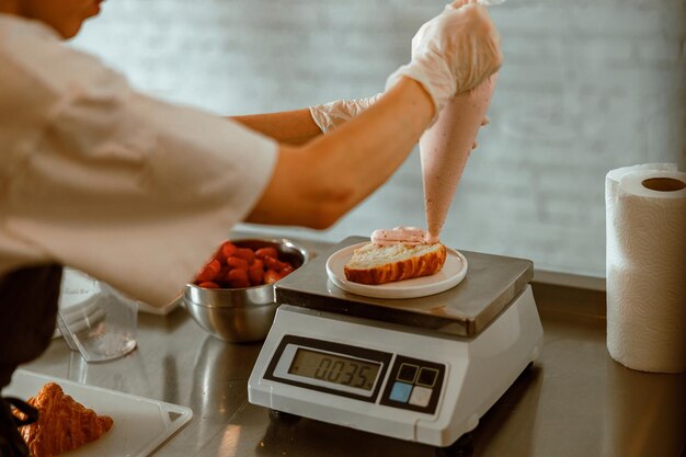 Confectioner applies pink cream onto croissant on scales in craft bakery shop