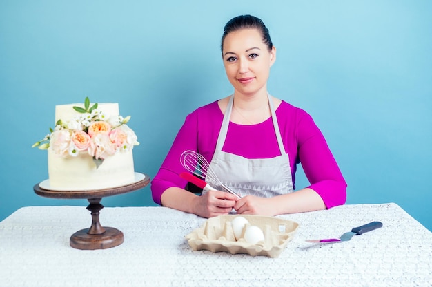 Confectioner housewife (pastry-cook) decorate appetizing creamy white two-tiered wedding cake with fresh flowers on a table with a lace tablecloth and holds a whisk in studio on a blue background