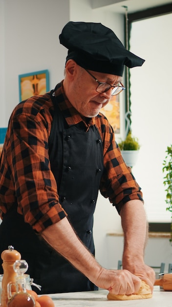 Confectioner working at home with raw dough in modern kitchen recording the recipe. Retired elderly baker with bonete mixing ingredients with sifted flour kneading for baking traditional bread.