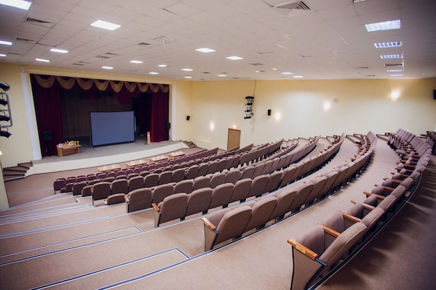 Foto sala riunioni per conferenze con luci a led a soffitto, sedie marrone fila, con palcoscenico e schermo vuoto per riunioni d'affari, conferenze