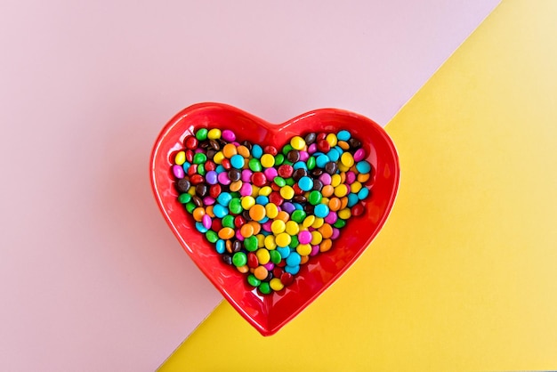 Confetti chocolates on heartshaped plate on yellow and pink background