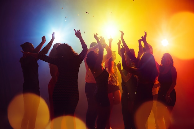 Confetti. A crowd of people in silhouette raises their hands on dancefloor on neon light background. Night life, club, music, dance, motion, youth. Yellow-blue colors and moving girls and boys.