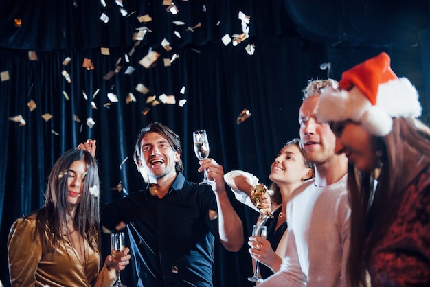 Confetti hangt in de lucht. Groep vrolijke vrienden die nieuwjaar binnenshuis vieren met drankjes in handen.