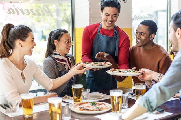 Confidant waiter serving delicious pizzas margherita to multicultural friends in cozy pizzeria restaurant Multiethnic friends having fun together at the pizzeria eating pizza and drinking blond beer