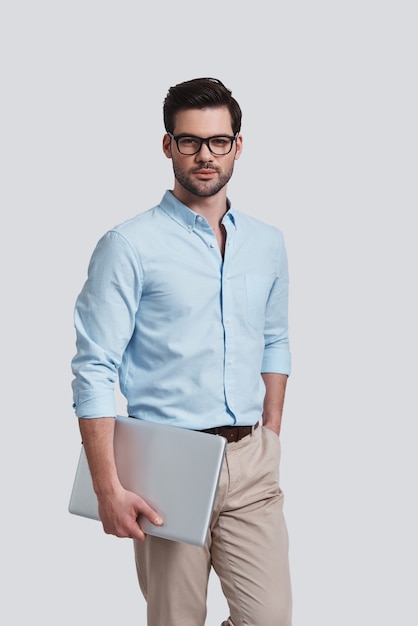 Confidence and charisma.  Good looking young man carrying laptop and looking at camera with smile while standing against grey background