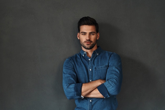 Confidence and charisma. Good looking young man keeping arms crossed and looking at camera while standing against grey background