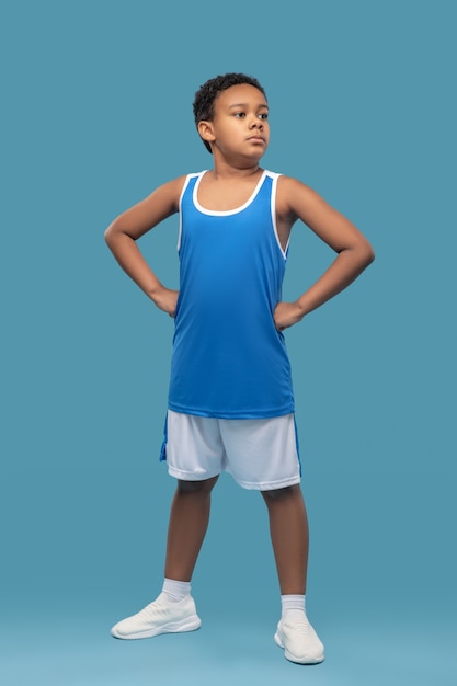 Уверенность. Темнокожий уверенный мальчик школьного возраста в спортивной одежде, стоя руки на поясе на синем фоне