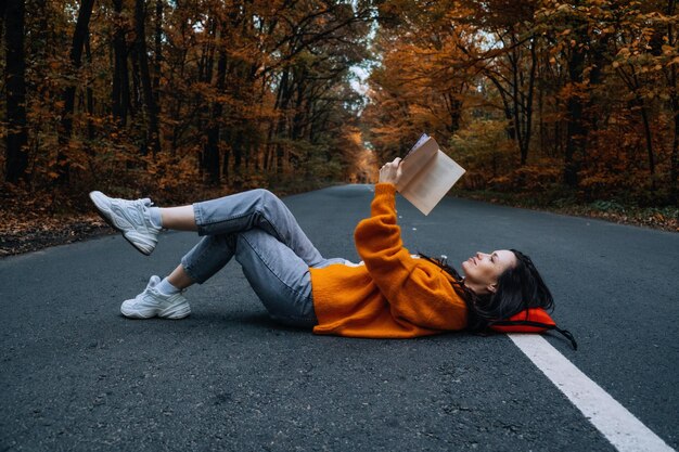 새로운 삶의 길을 선택하는 가을 나무와 함께 도로에 앉아있는 책과 함께 자신감있는 여성