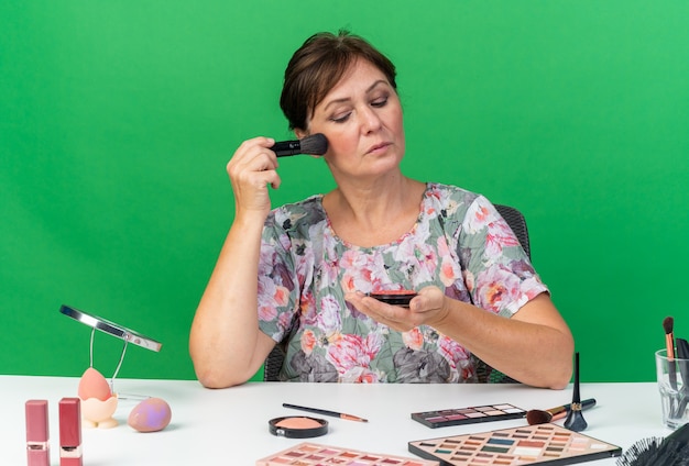 Confident adult caucasian woman sitting at table with makeup tools holding and looking at blush applying with makeup brush 